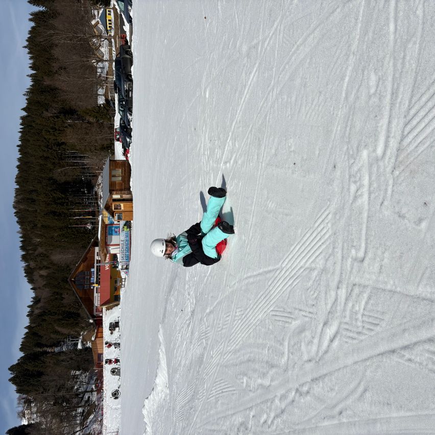 Ein Snowboarder liegt auf dem Rücken auf einem schneebedeckten Hang mit Spuren, trägt einen Helm und eine blaue Jacke. Hinter ihm stehen Gebäude, Bäume und geparkte Autos.