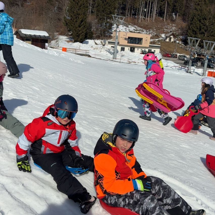 Mehrere Kinder sitzen auf Schneeschlitten auf einem verschneiten Hügel. Sie tragen Helme und farbenfrohe Winterjacken. Eine Mädchen läuft mit einem rosa Schlitten hinter ihnen. Im Hintergrund sind Bäume, ein Gebäude und geparkte Autos zu sehen.