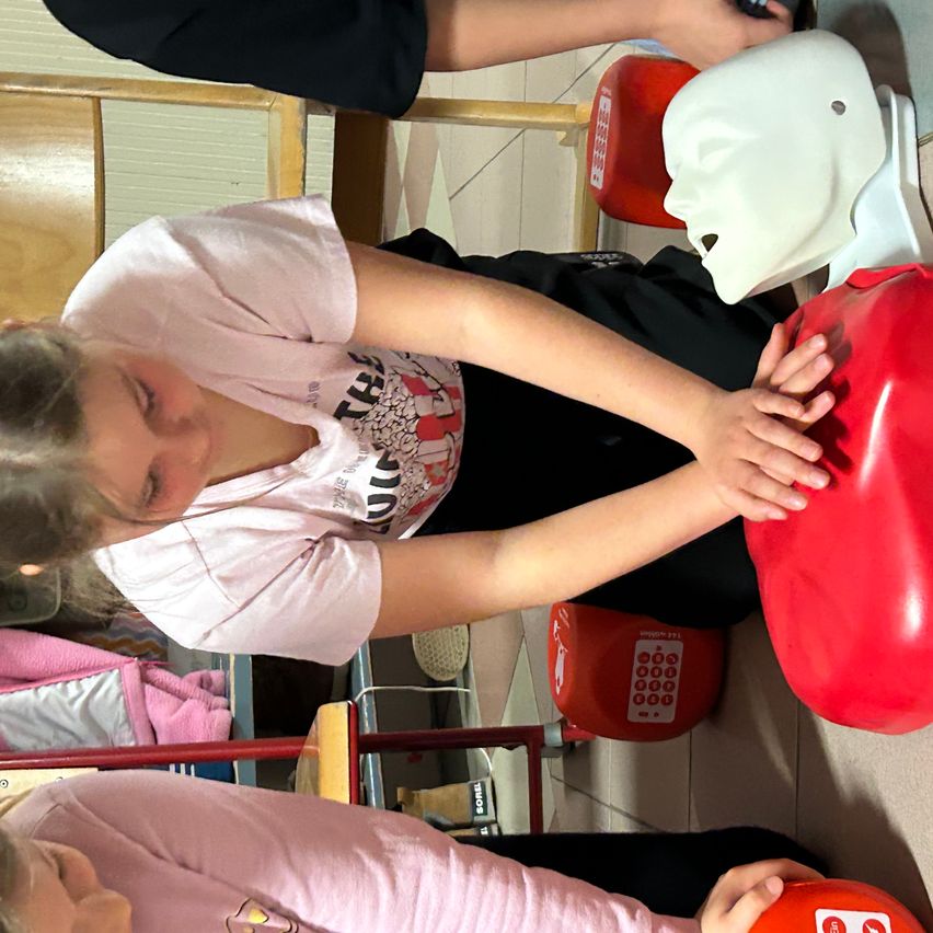 Ein Mädchen übt CPR an einem roten Brustmannequin. Hinter ihr hält eine weitere Person ein Telefon. In der Nähe ist ein roter Notrufkasten an der Wand angebracht.