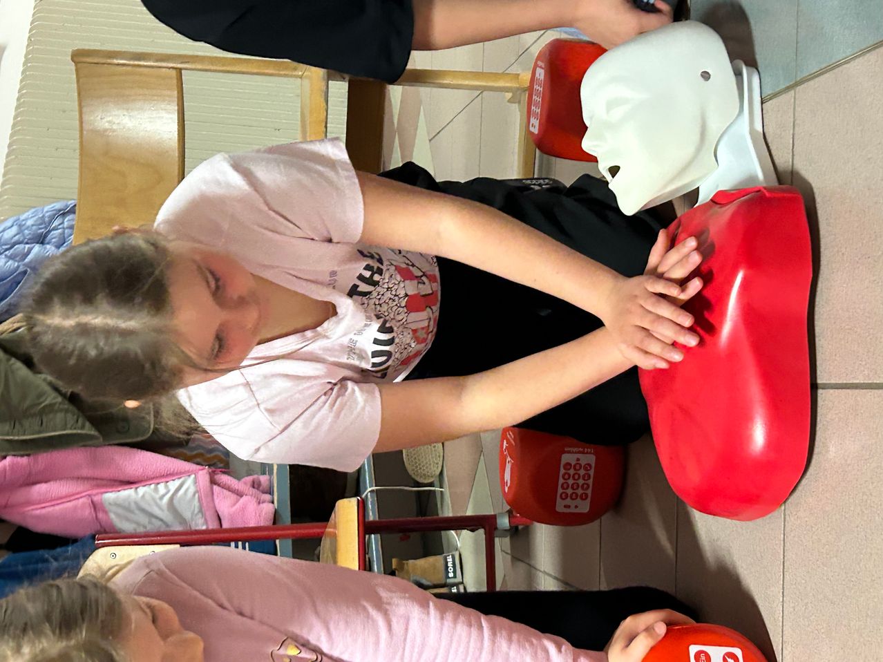 Ein Mädchen übt CPR an einem roten Brustmannequin. Hinter ihr hält eine weitere Person ein Telefon. In der Nähe ist ein roter Notrufkasten an der Wand angebracht.