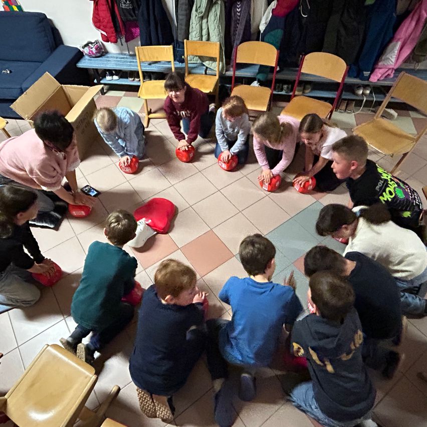 Eine Gruppe von Kindern übt CPR mit Dummies auf dem Boden, angeleitet von Erwachsenen. Der Raum hat Stühle und Jacken auf einer nahen Bank.