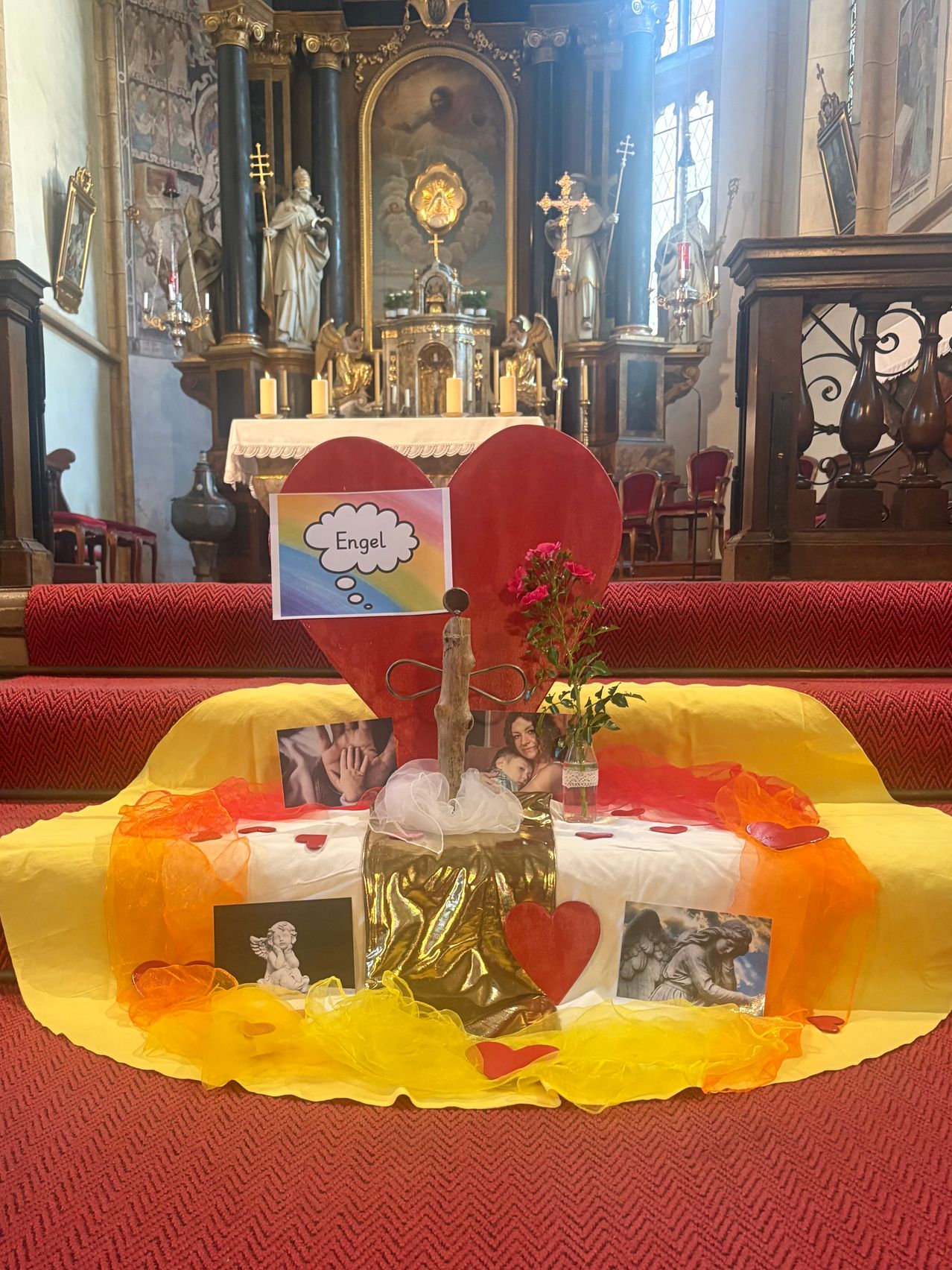 Ein herzförmiger Altar in einer Kirche mit einem Schild, das Engel steht, umgeben von Fotos, Blumen und dekorativen Elementen.