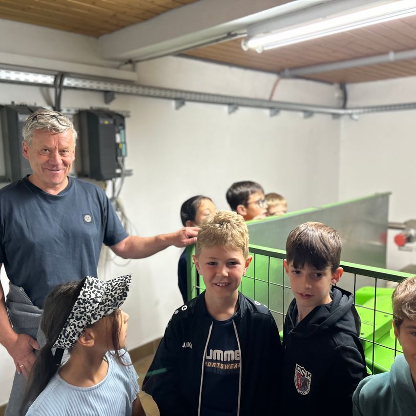 Eine Gruppe von Kindern und ein Mann lächeln in einem Raum, wobei ein Junge von dem Mann auf den Kopf tätschelt. Sie stehen vor einem grünen Metallzaun.