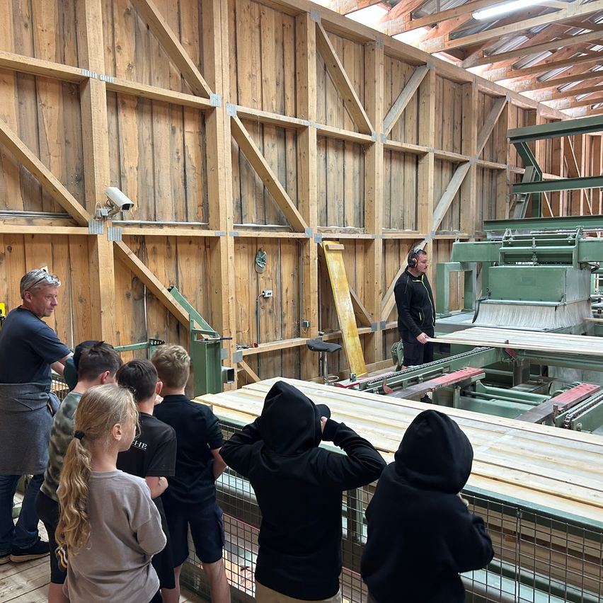 Eine Gruppe von Menschen befindet sich in einer Fabrik und beobachtet einen Mann, der eine große Maschine bedient. Die Wände bestehen aus Holz und Metall.