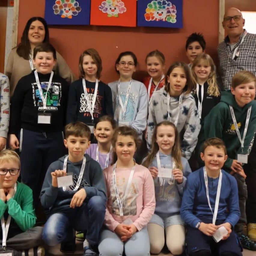 Eine Gruppe von Kindern mit Namensschildern posiert für ein Foto mit zwei Erwachsenen. Die Kinder sitzen und knien auf der Bühne. An der Wand hinter ihnen hängen bunte Gemälde.