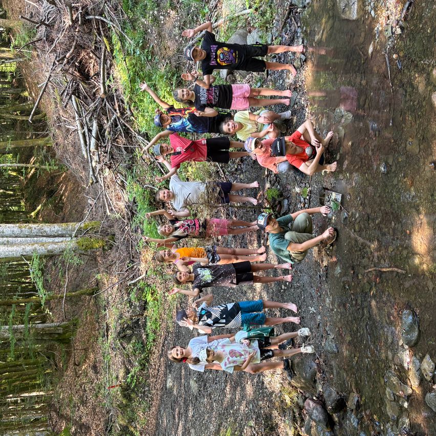 Eine Gruppe junger Leute versammelt sich in einem Wald, einige posieren für ein Foto an einem Bach. Sie befinden sich in verschiedenen Positionen, wobei sich einige gegen die Felsen lehnen.