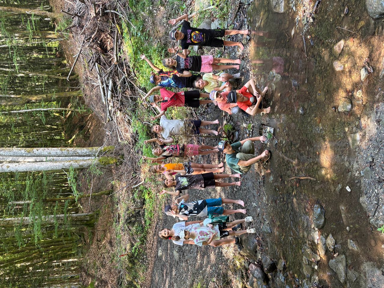 Eine Gruppe junger Leute versammelt sich in einem Wald, einige posieren für ein Foto an einem Bach. Sie befinden sich in verschiedenen Positionen, wobei sich einige gegen die Felsen lehnen.