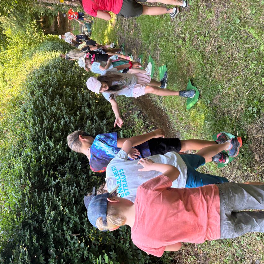 Eine Gruppe von Kindern in verschiedenen Farben von Shirts und Hosen steht in einer Reihe in einem Grasfeld. Einige tragen Hüte und andere haben Turnschuhe an. Sie scheinen an einer Aktivität teilzunehmen.
