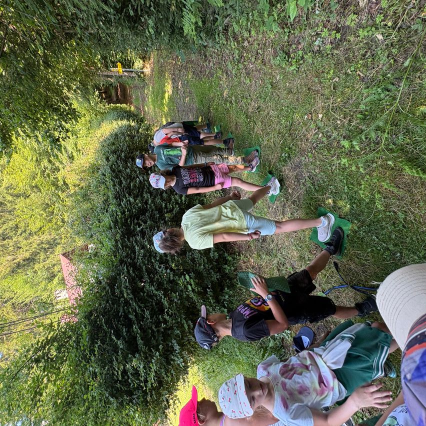 Eine Gruppe von Kindern liegt auf dem Gras in einem Wald, einige tragen Mützen und Turnschuhe. Sie scheinen die Umgebung im Freien zu genießen.