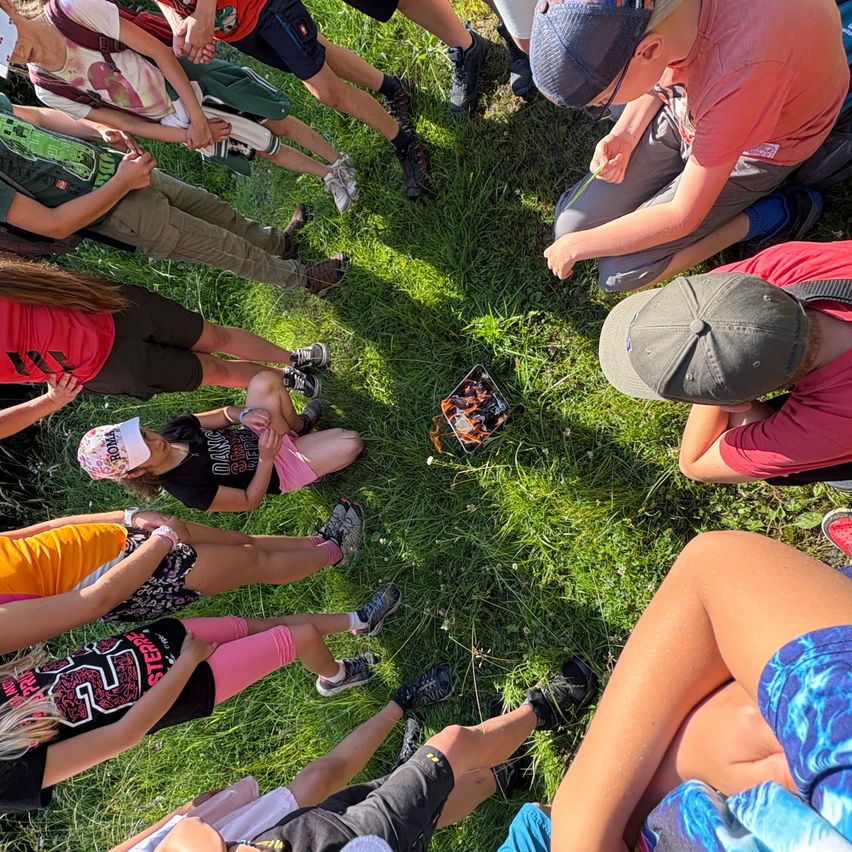 Eine Gruppe von Menschen sitzt auf dem Gras, einige tragen Hüte und Sneakers. Eine Person hält eine Schachtel mit Flammendesign.