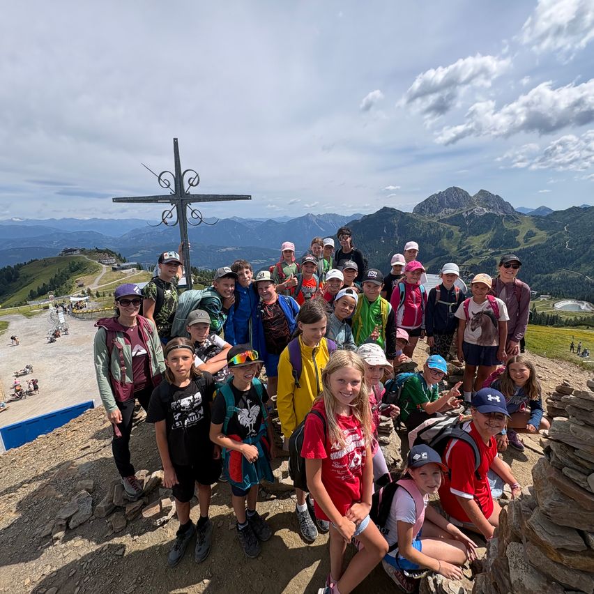 Eine Gruppe von Menschen posiert für ein Foto auf einem Berg mit einem Kreuz im Hintergrund. Sie tragen Hüte und Rucksäcke und lächeln für die Kamera.