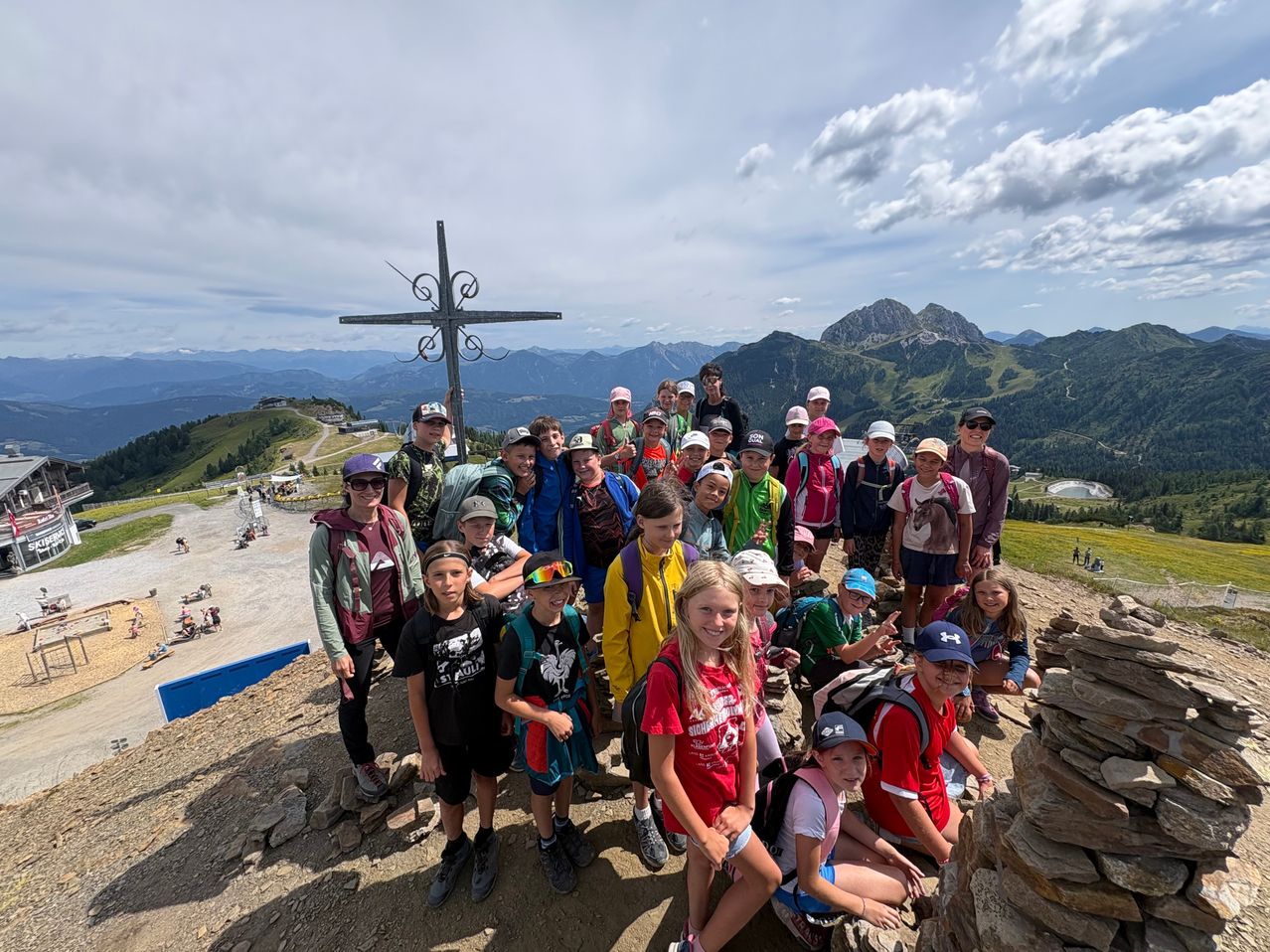 Eine Gruppe von Menschen posiert für ein Foto auf einem Berg mit einem Kreuz im Hintergrund. Sie tragen Hüte und Rucksäcke und lächeln für die Kamera.
