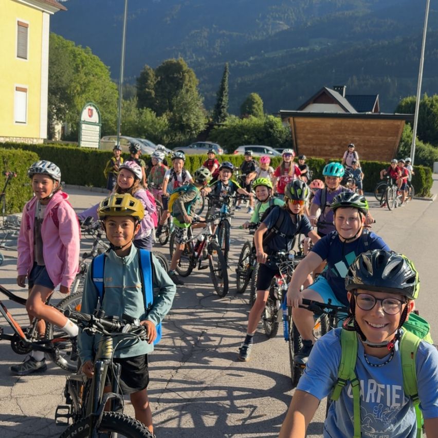 Eine Gruppe von Kindern mit Helmen und Rucksäcken steht mit ihren Fahrrädern in einem Park. Sie stehen auf einer Straße mit Bergen im Hintergrund.