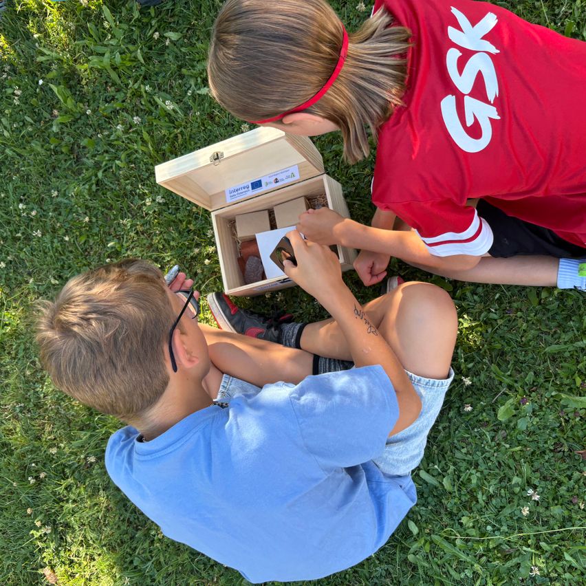 Zwei Kinder untersuchen eine kleine Holzbox mit einer Karte darin, eines in einem roten GSK-Shirt und das andere in Blau, auf einem Grasfeld sitzend.