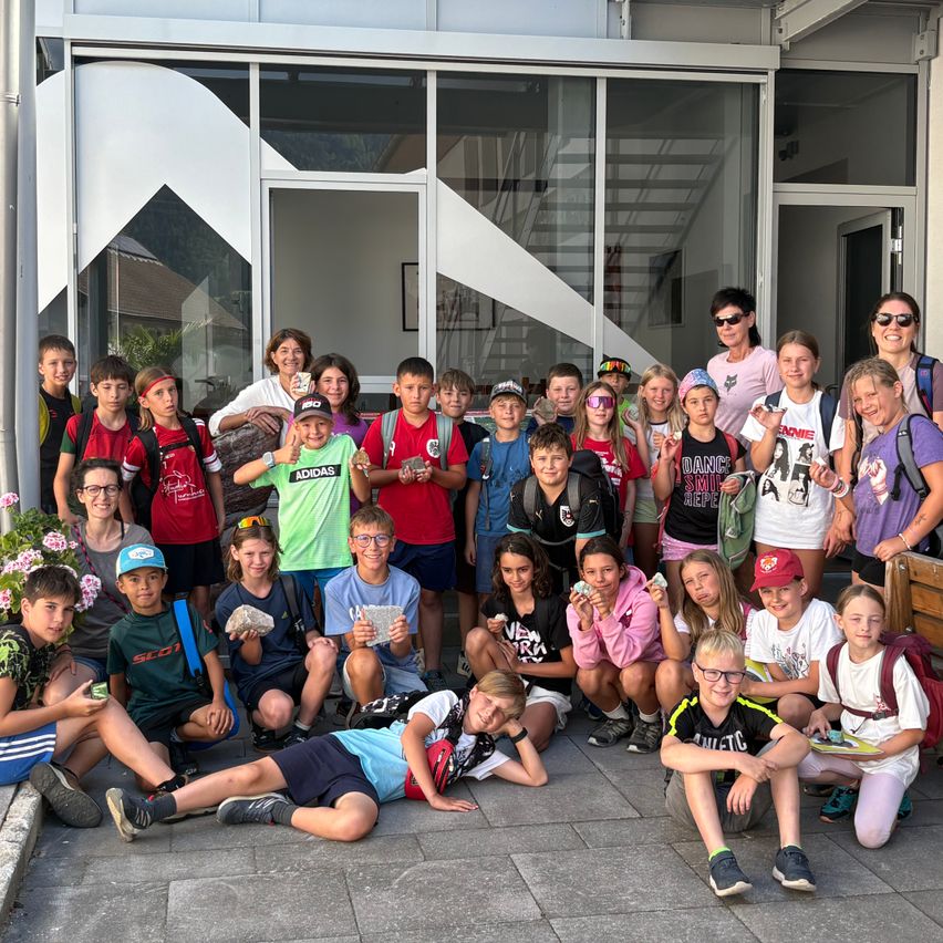 Eine Gruppe von Schülern, einige mit Steinen in der Hand, posiert für ein Foto vor einem Gebäude. Sie tragen verschiedene farbige Shirts und Mützen. Zwei Frauen stehen hinter ihnen und lächeln.