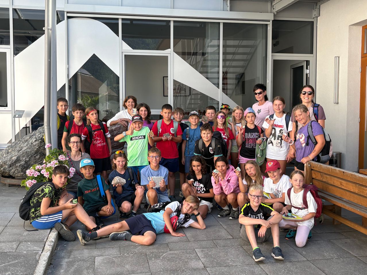Eine Gruppe von Schülern, einige mit Steinen in der Hand, posiert für ein Foto vor einem Gebäude. Sie tragen verschiedene farbige Shirts und Mützen. Zwei Frauen stehen hinter ihnen und lächeln.