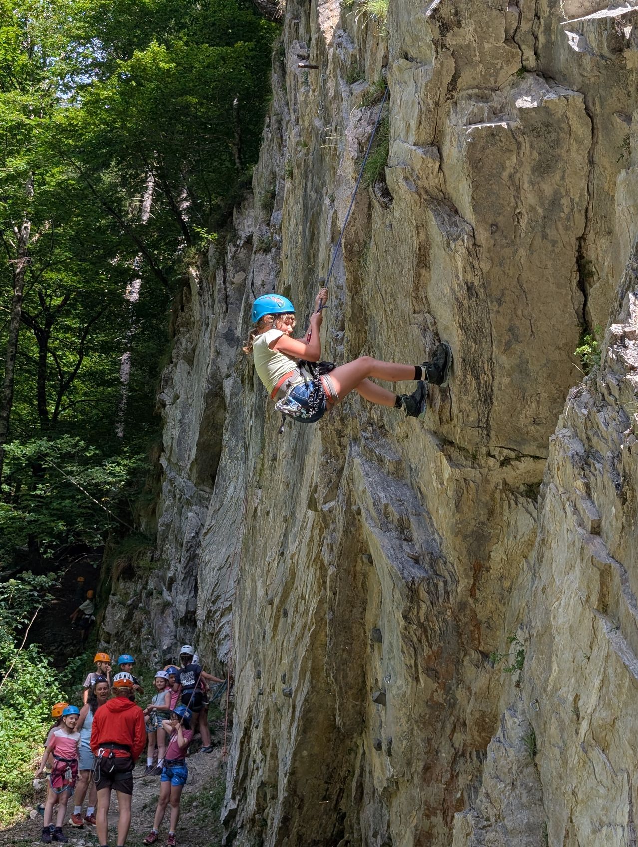 Ein junges Mädchen mit blauem Helm klettert an einer Felswand mit einem Seil, während andere von unten zusehen.
