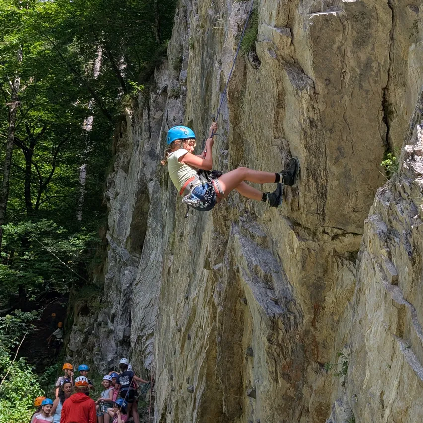 Ein junges Mädchen mit blauem Helm klettert an einer Felswand mit einem Seil, während andere von unten zusehen.