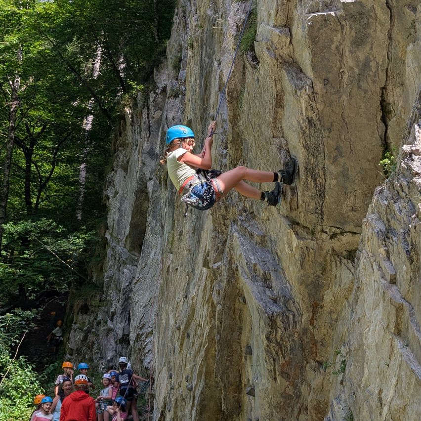 Ein junges Mädchen mit blauem Helm klettert an einer Felswand mit einem Seil, während andere von unten zusehen.