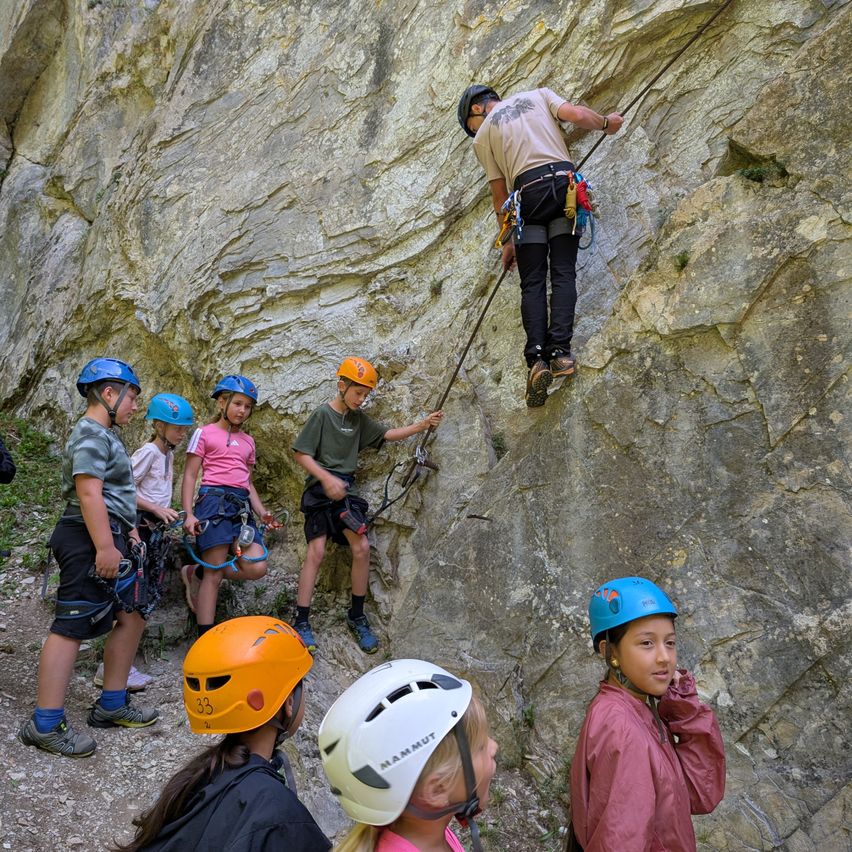 Eine Gruppe von Kindern in Helmen beobachtet einen Mann, der mit einem Seil eine Felswand erklimmt.