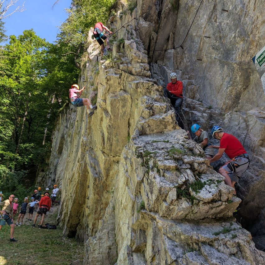 Eine Gruppe von Menschen klettert auf einem steilen Felsvorsprung, einige tragen Helme und Gurte. Bäume und ein blaues Schild sind im Hintergrund sichtbar.