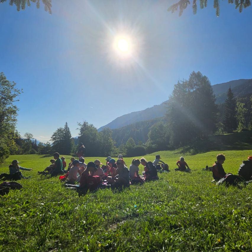 Eine Gruppe von Menschen sitzt auf einer grasbewachsenen Wiese unter einem hellen Sonnenschein, umgeben von Bäumen und Bergen.