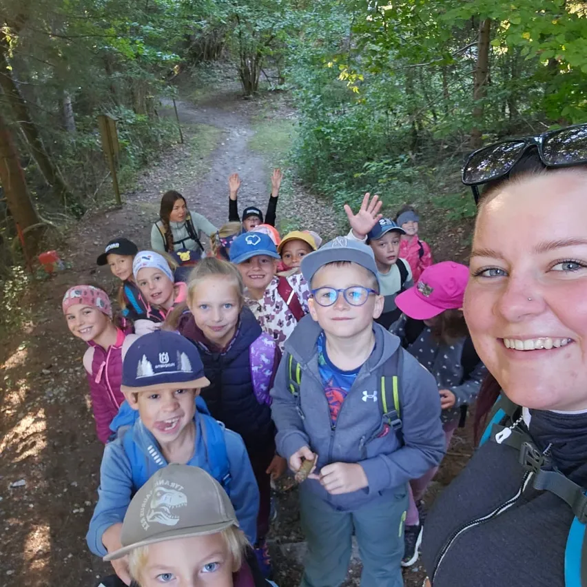 Eine Gruppe von Kindern und Erwachsenen auf einem Waldweg, sie lächeln und winken, mit einer Frau im Vordergrund.
