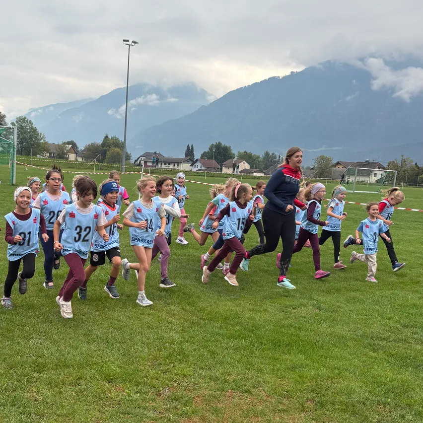 Eine Gruppe von Kindern in blauen Trikots mit Nummern läuft auf einem Rasenfeld. Eine Frau in einer roten Jacke führt sie an. Im Hintergrund befinden sich Häuser und Berge.
