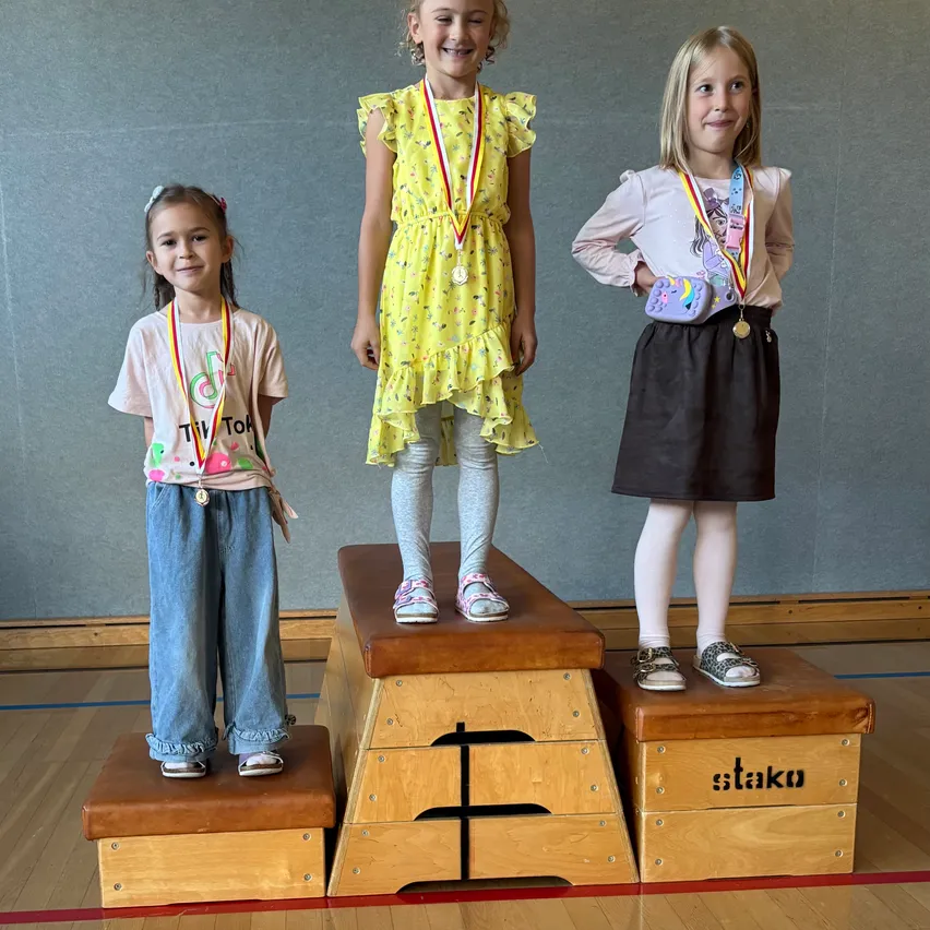 Drei junge Mädchen stehen auf einer Holzplattform, wahrscheinlich bei einer Schulveranstaltung. Das Mädchen in der Mitte trägt ein gelbes Kleid und hat eine Medaille um den Hals. Das Mädchen auf der linken Seite trägt ein rosa Shirt und hat eine Medaille um den Hals. Das Mädchen auf der rechten Seite trägt ein weißes Shirt und hat eine Medaille um den Hals.