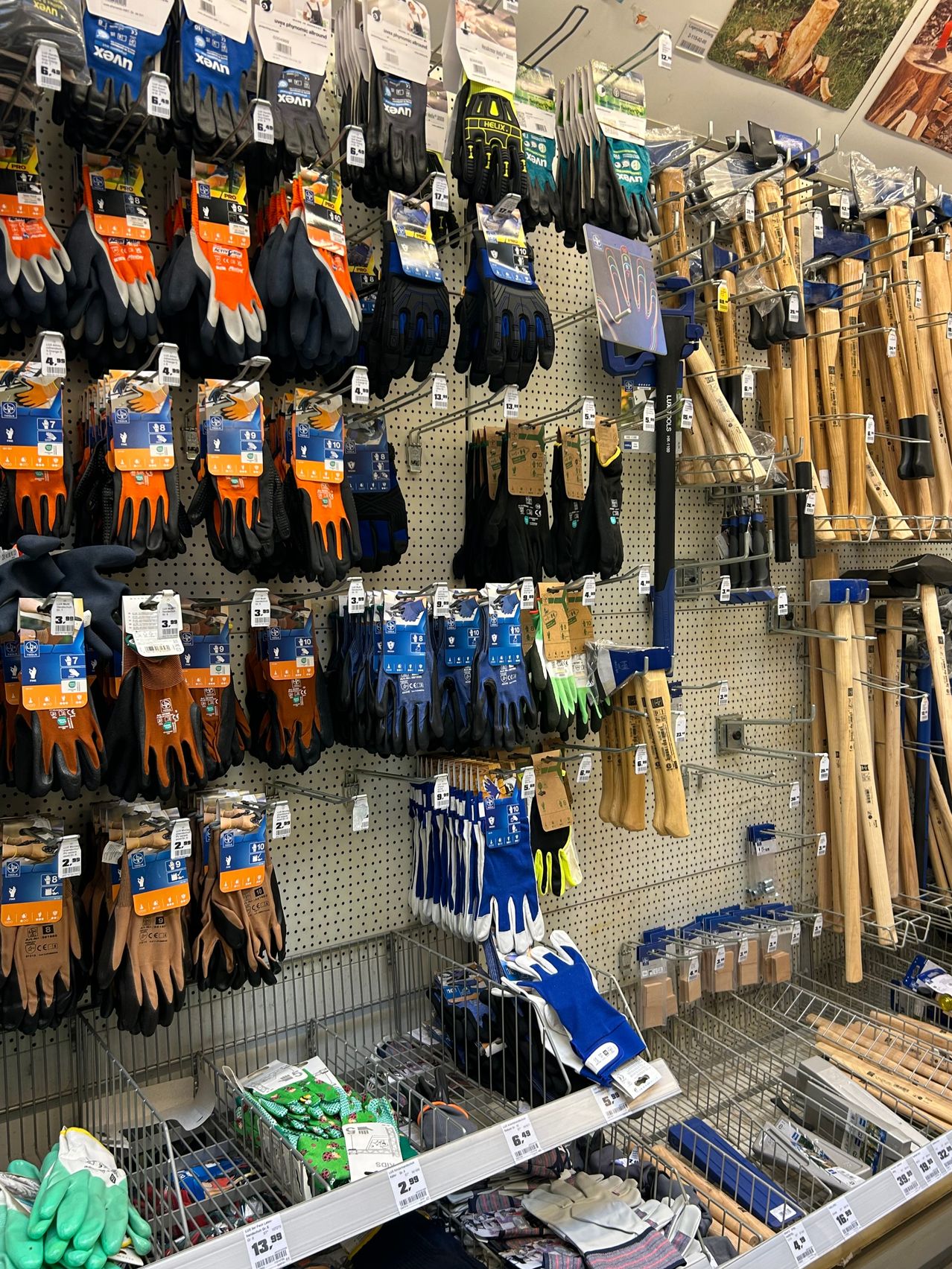 Ein Geschäft zeigt verschiedene Handschuhe und Werkzeuge, einschließlich orangefarbener, schwarzer und blauer Handschuhe, mit hölzernen und metallischen Werkzeugen, die an der Wand hängen.