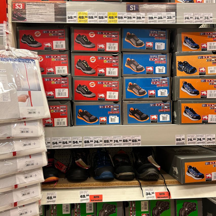 Ein Supermarktregal zeigt mehrere Paare von Sicherheitsschuhen in verschiedenen Farben. Einige Schuhe sind in ihren Schachteln, während andere direkt auf den Regalen platziert sind. Die Preisschilder sind an den Schachteln und Schuhen sichtbar.