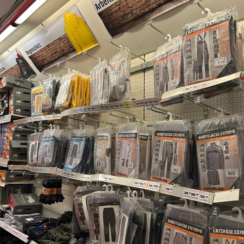 Ein Einzelhandelsgeschäft zeigt eine Vielzahl von Arbeitskleidung, einschließlich Hosen, Jacken und Handschuhen, alle ordentlich verpackt. Über den Produkten werben Banner mit 'Arbeitsschutz'. Das Geschäft hat weiße Wände und Deckenleuchten.