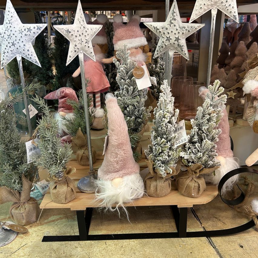 Eine Ausstellung von Weihnachtsdekorationen umfasst kleine schneebedeckte Bäume, einen Gnom und rosa Sterne. Die Dekorationen sind in Jute gewickelt und auf einem Schlitten platziert.