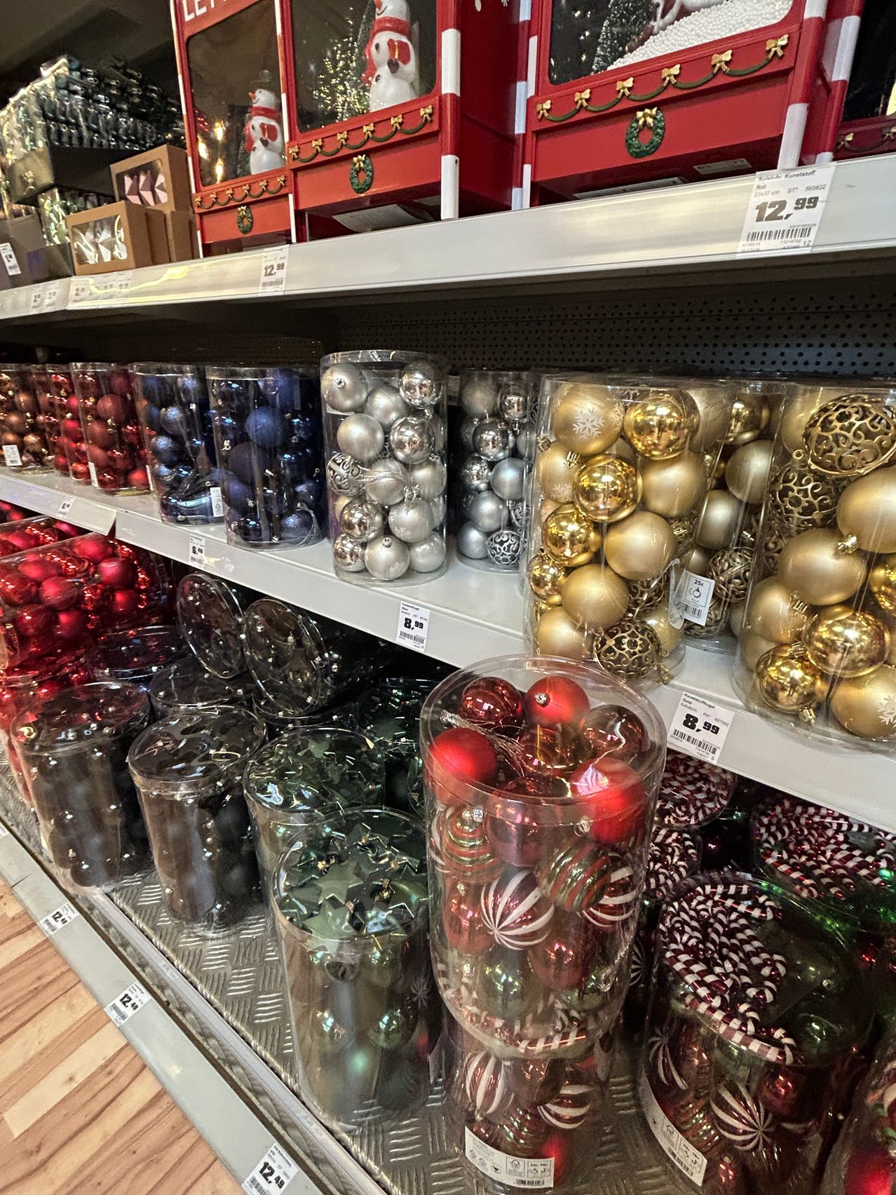 Ein Ladenregal gefüllt mit verschiedenen durchsichtigen Behältern, die Weihnachtskugeln enthalten. Die Behälter sind in verschiedenen Farben, wie rot, gold, silber und blau. Die Behälter sind ordentlich in Reihen angeordnet. Die Preisschilder sind an den Behältern befestigt.