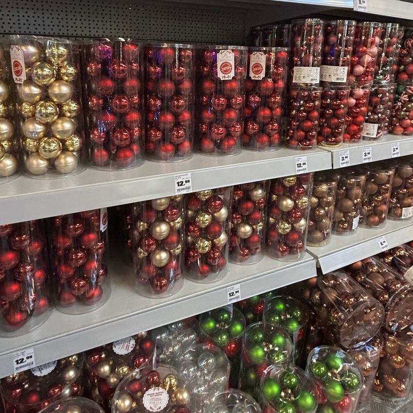 Ein Ladenregal zeigt verschiedene durchsichtige Kunststoffbehälter, die mit roten und goldenen Weihnachtskugeln gefüllt sind. Mehrere Reihen von Behältern sind gestapelt. Einige Behälter enthalten auch grüne Kugeln.