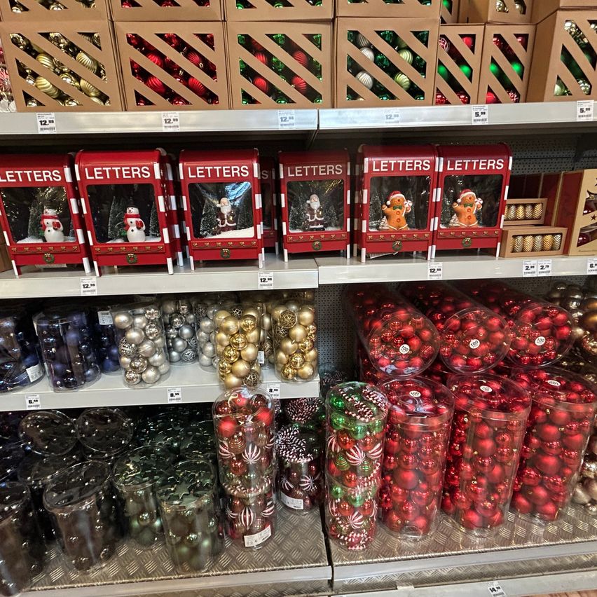 Ein Geschäft zeigt eine Vielzahl von Weihnachtsschmuck, darunter rote und grüne Kugeln, durchsichtige Kunststoffbehälter mit Kugeln und rote Geschenkboxen mit Lebkuchenmännern auf der Oberseite. Die Regale sind mit einer Auswahl an Ornamenten in verschiedenen Größen und Farben gefüllt.