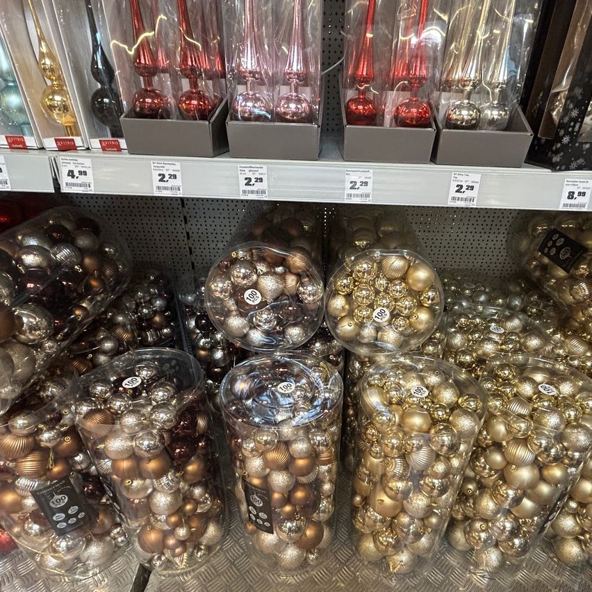 Ein Ladenregal mit mehreren transparenten Behältern, die verschiedene goldene und silberne Weihnachtskugeln enthalten. Die Behälter sind nummeriert und bepreist. Über den Behältern sind Schachteln mit roten und silbernen Ornamenten auf einem Regal ausgestellt.