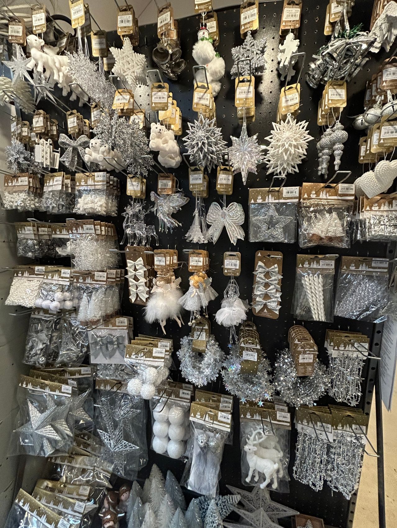 Ein Ladenregal mit verschiedenen Weihnachtsdekorationen, darunter Schneeflocken, Engelsfiguren, Schleifen, Tinsel und silberne Ornamente. Viele Artikel sind in Plastikverpackungen mit Etiketten.