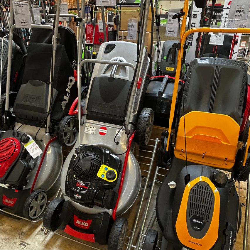Mehrere Rasenmäher in verschiedenen Farben sind in einem Geschäft ausgestellt. Die Mäher haben verschiedene Marken, darunter AL-KO und Honda. Die Mäher haben verschiedene Funktionen, wie einen schwarzen Motor und einen gelben Griff.