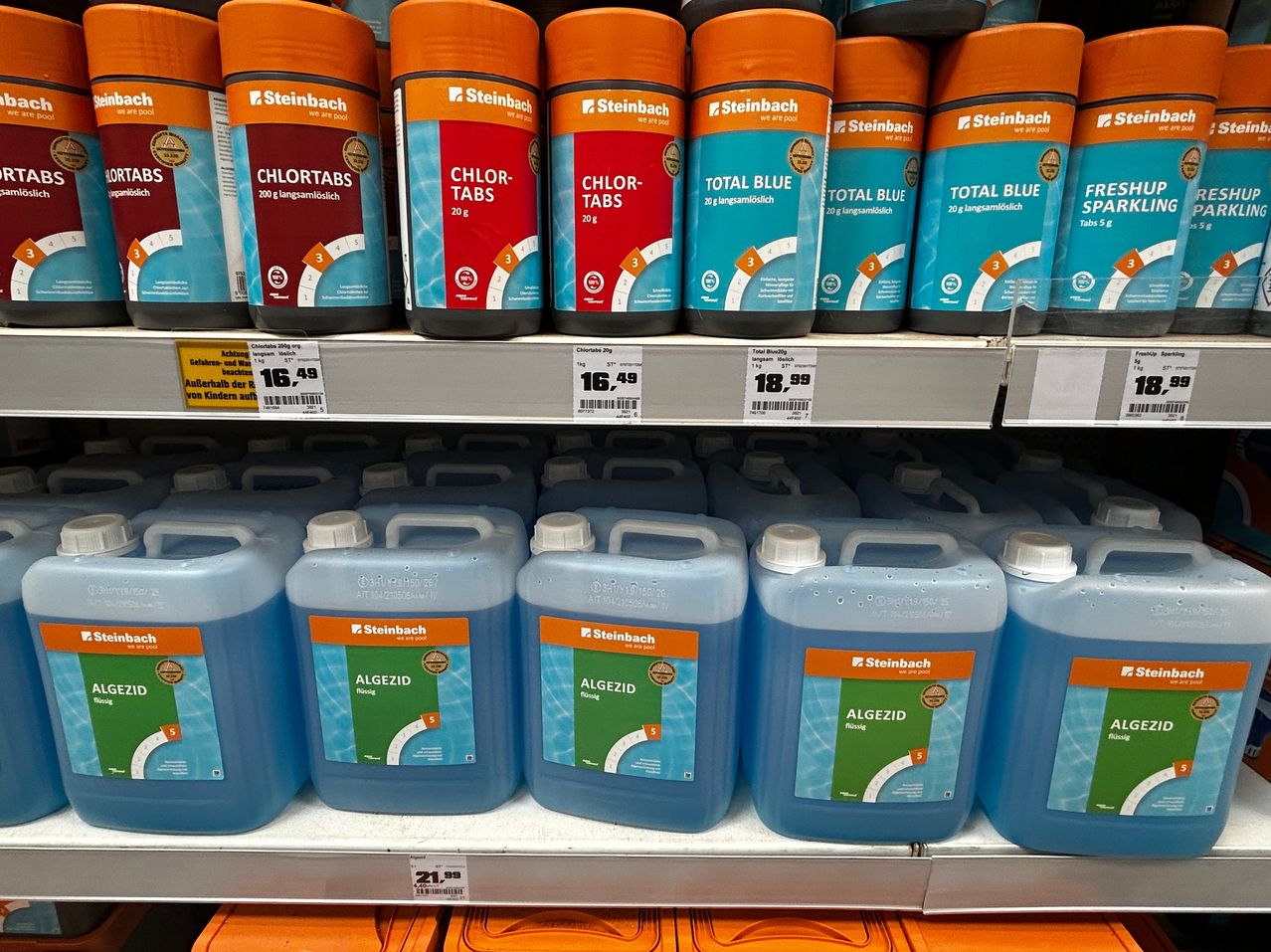 A store shelf displays several bottles of pool chemicals, including Chlor-Tabs and Algezide, with price tags visible. The bottles are neatly arranged in rows, showcasing their vibrant colors and labels.