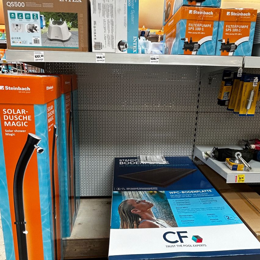 A store shelf displays various pool products, including boxes of solar-powered showers, filter pumps, and a floor mat. The boxes are prominently labeled with their names and prices.