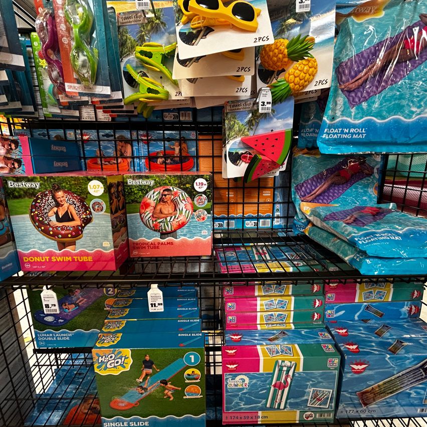 A store shelf displays various pool floats and inflatable toys, including a donut swim tube, tropical palms swim tube, and a single slide. The products are neatly arranged on black shelves.