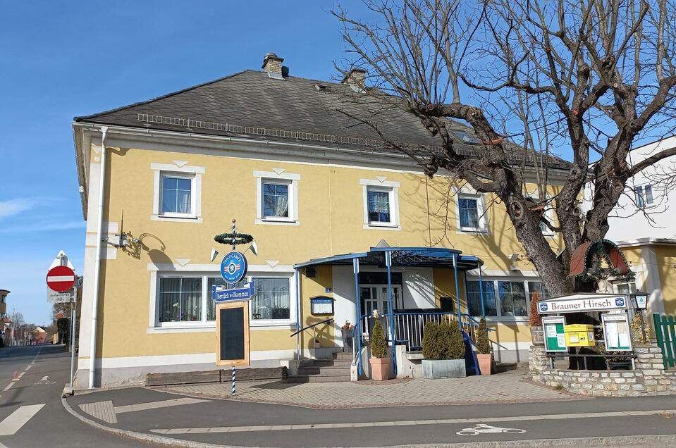 Ein gelbes Gebäude mit mehreren Fenstern und einem blauen Vordach. Ein Kreidetafelschild steht davor. Ein großer Baum steht neben dem Gebäude.