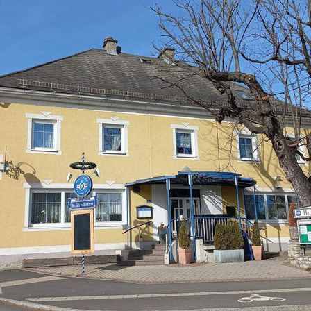 Ein gelbes Gebäude mit mehreren Fenstern und einem blauen Vordach. Ein Kreidetafelschild steht davor. Ein großer Baum steht neben dem Gebäude.
