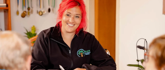 Eine Frau mit roten Haaren, die eine schwarze Jacke mit Logo trägt, sitzt an einem Tisch, lächelt und schreibt in ein Notizbuch. Ein Glas Wasser und ein Stift sind auf dem Tisch. Eine andere Person sitzt neben ihr.