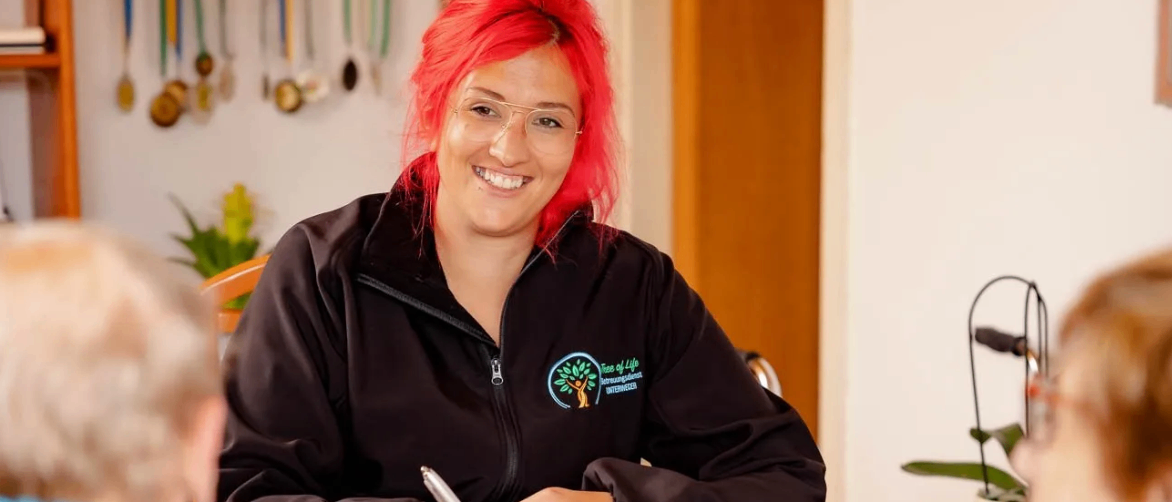 Eine Frau mit roten Haaren, die eine schwarze Jacke mit Logo trägt, sitzt an einem Tisch, lächelt und schreibt in ein Notizbuch. Ein Glas Wasser und ein Stift sind auf dem Tisch. Eine andere Person sitzt neben ihr.