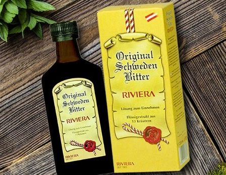 Eine Flasche Original Schweden Bitter liegt neben ihrer gelben Schachtel auf einer Holzoberfläche, umgeben von grünen Pflanzen.