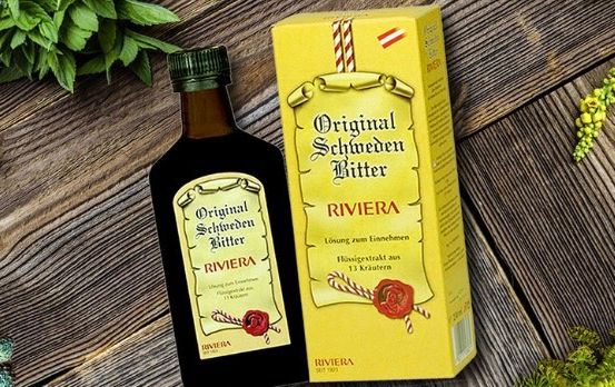 Eine Flasche Original Schweden Bitter liegt neben ihrer gelben Schachtel auf einer Holzoberfläche, umgeben von grünen Pflanzen.