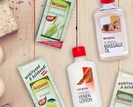 Auf einem Holztisch sind verschiedene Hautpflegeprodukte ausgestellt. Zu den Produkten gehören eine Flasche Riviera Massageöl, zwei Flaschen Riviera Venen Lotion und einige Beutel pur Lemongrass und Grapefruit Saunaöl. Es gibt auch zwei weiße Kerzen.