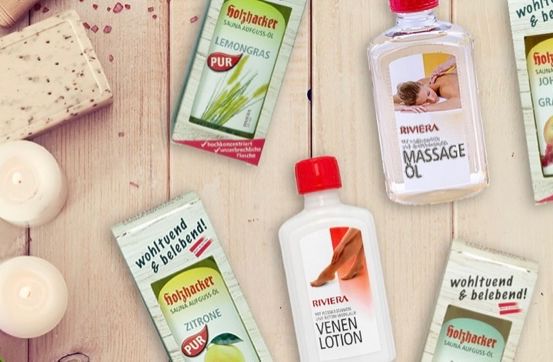 Auf einem Holztisch sind verschiedene Hautpflegeprodukte ausgestellt. Zu den Produkten gehören eine Flasche Riviera Massageöl, zwei Flaschen Riviera Venen Lotion und einige Beutel pur Lemongrass und Grapefruit Saunaöl. Es gibt auch zwei weiße Kerzen.